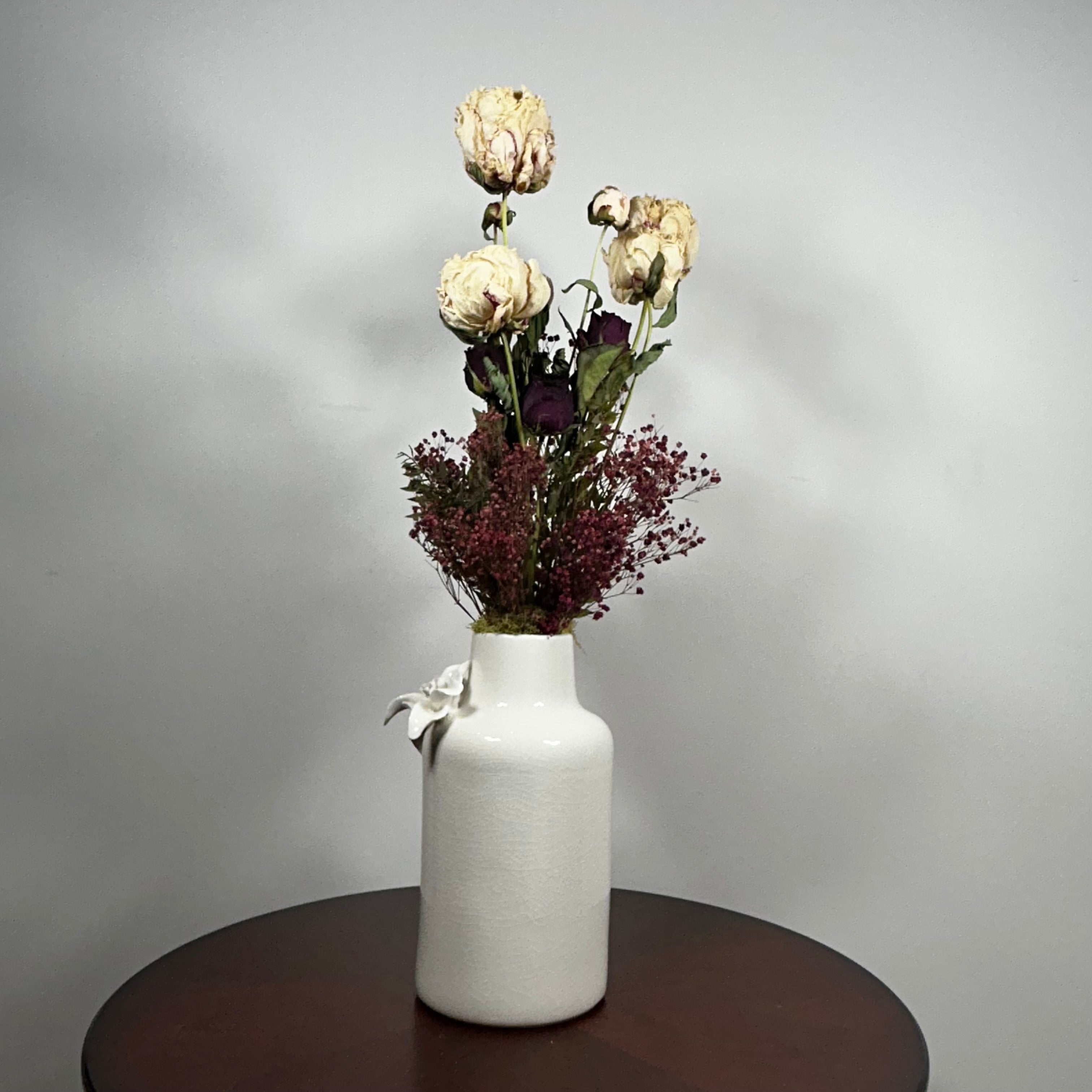 A white ceramic vase with a textured finish, decorated with a small white bow, holding a dried flower arrangement with cream and burgundy tones, set on a dark round table against a light gray wall.