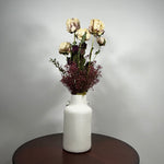 A white ceramic vase with small side handles, filled with dried cream-colored flowers and burgundy filler stems, set on a dark round wooden table against a light gray wall.