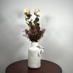 A white textured ceramic vase with a white ribbon bow, filled with dried cream and burgundy flowers, sitting on a dark round wooden table against a light gray wall.