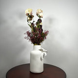 A white textured ceramic vase with a white ribbon bow, filled with dried cream and burgundy flowers, sitting on a dark round wooden table against a light gray wall.