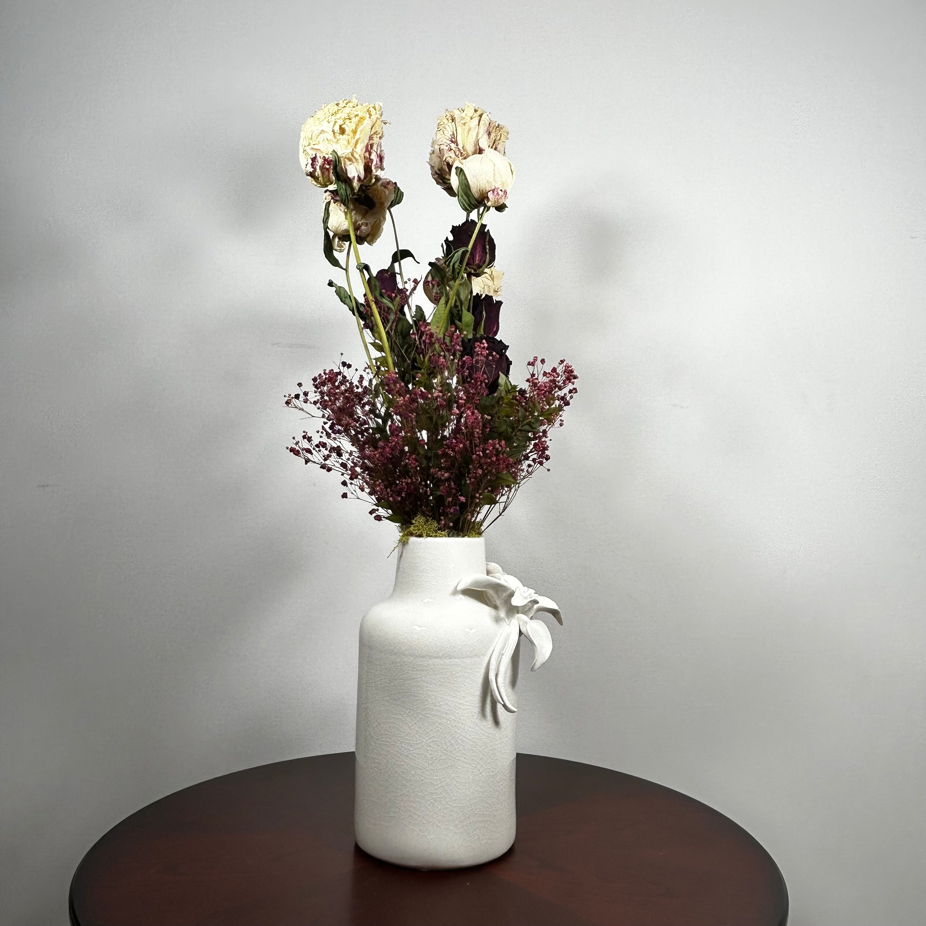 A white textured ceramic vase with a white ribbon bow, filled with dried cream and burgundy flowers, sitting on a dark round wooden table against a light gray wall.