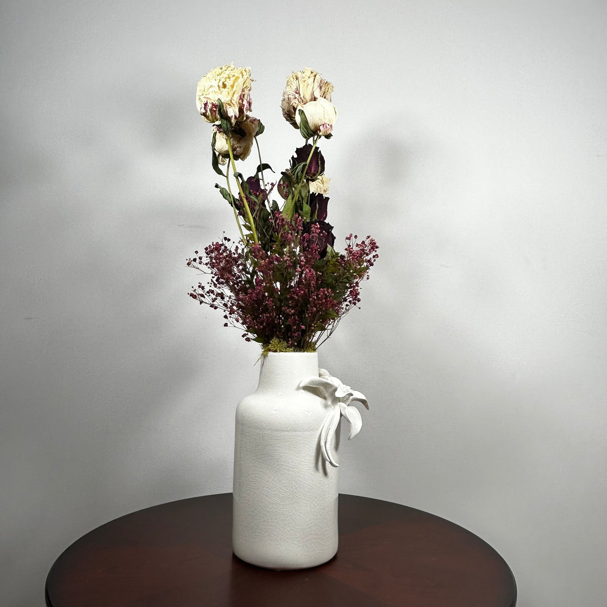 A white textured ceramic vase with a white ribbon bow, filled with dried cream and burgundy flowers, sitting on a dark round wooden table against a light gray wall.