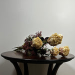 A dried floral arrangement with cream roses, burgundy filler flowers, and dark green stems resting on a dark wooden table against a plain light gray wall.
