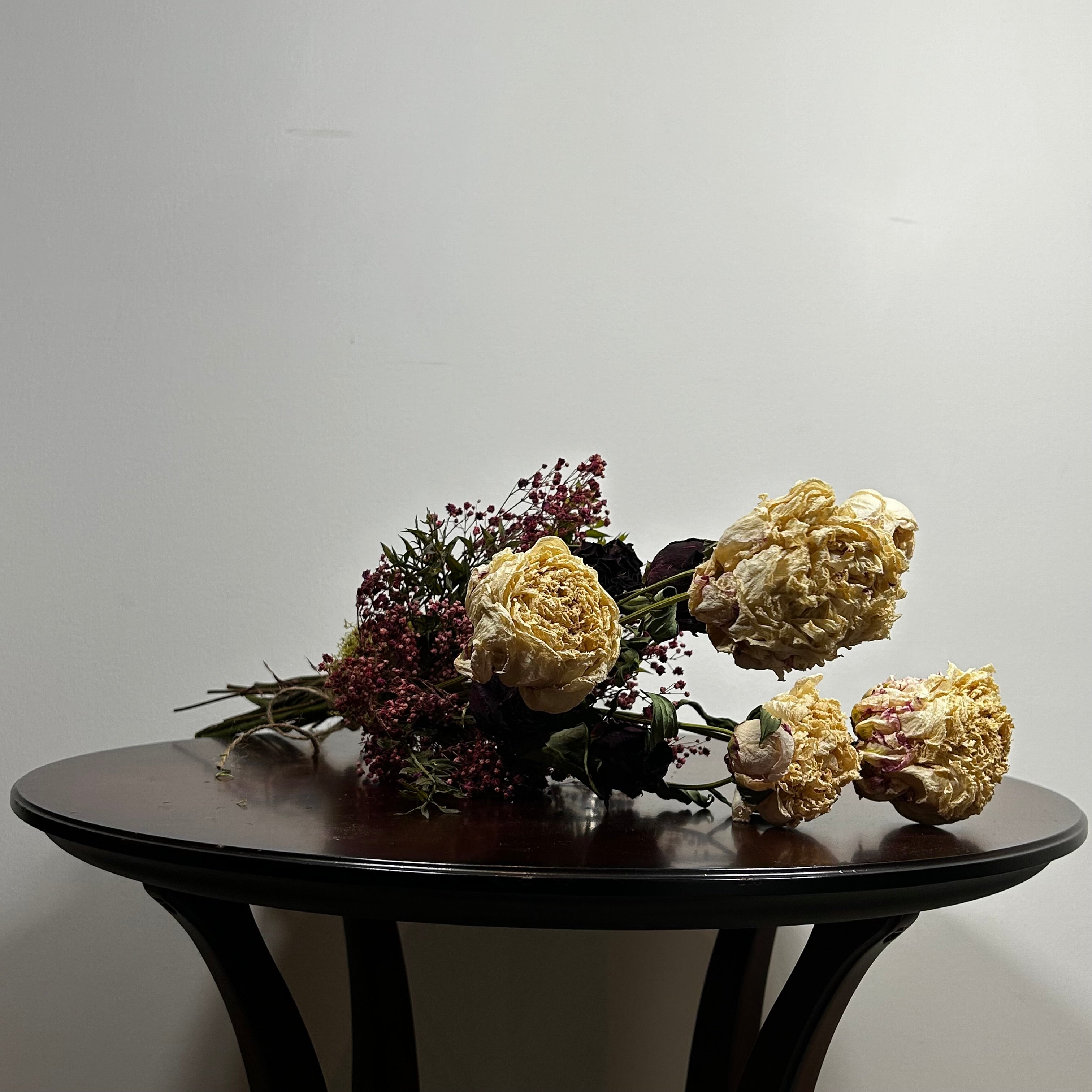 A dried floral arrangement with cream roses, burgundy filler flowers, and dark green stems resting on a dark wooden table against a plain light gray wall.
