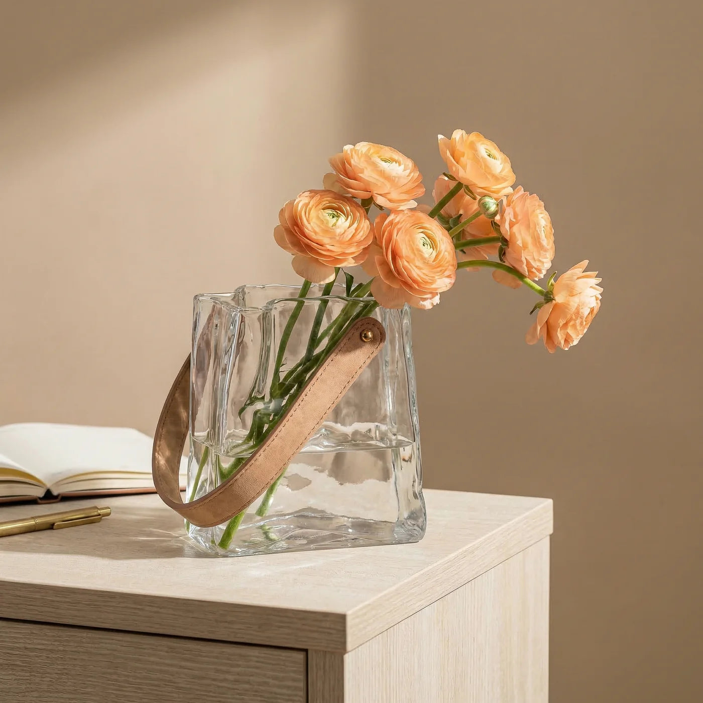 Clear glass vase shaped like a paper bag with faux leather handle holding peach-coloured flowers on a wooden surface with a beige background.