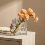 Clear glass vase shaped like a paper bag with faux leather handle holding peach-coloured flowers on a wooden surface with a beige background.