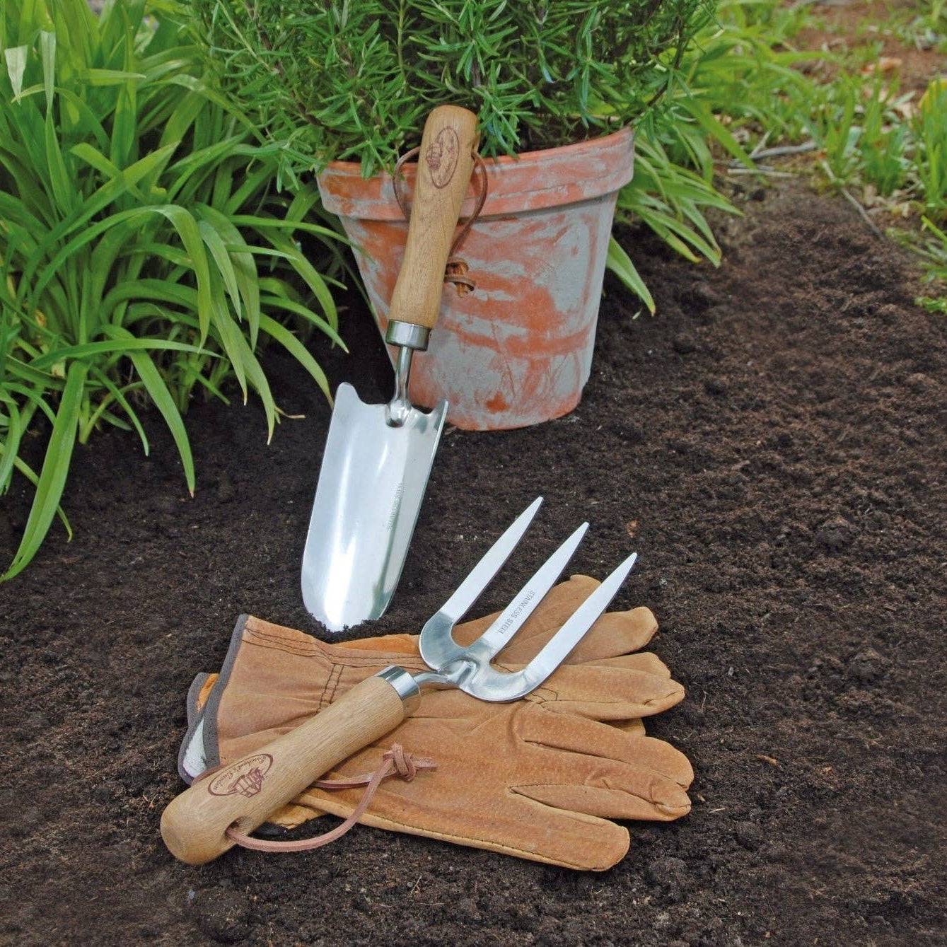 Gardening tools including a trowel, fork, and gloves on garden soil with a potted plant in the background.