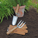 Gardening tools including a trowel, fork, and gloves on garden soil with a potted plant in the background.