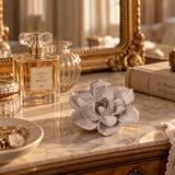 A decorative gray ceramic flower sculpture sits on a marble vanity table alongside a glass perfume bottle, a gold mirror, a vintage book, and a lace cloth in a warm, elegant interior setting.