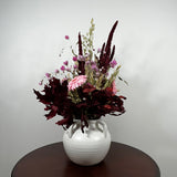 A dried flower arrangement in a white ceramic vase with two handles, set on a dark round table against a plain light gray wall.