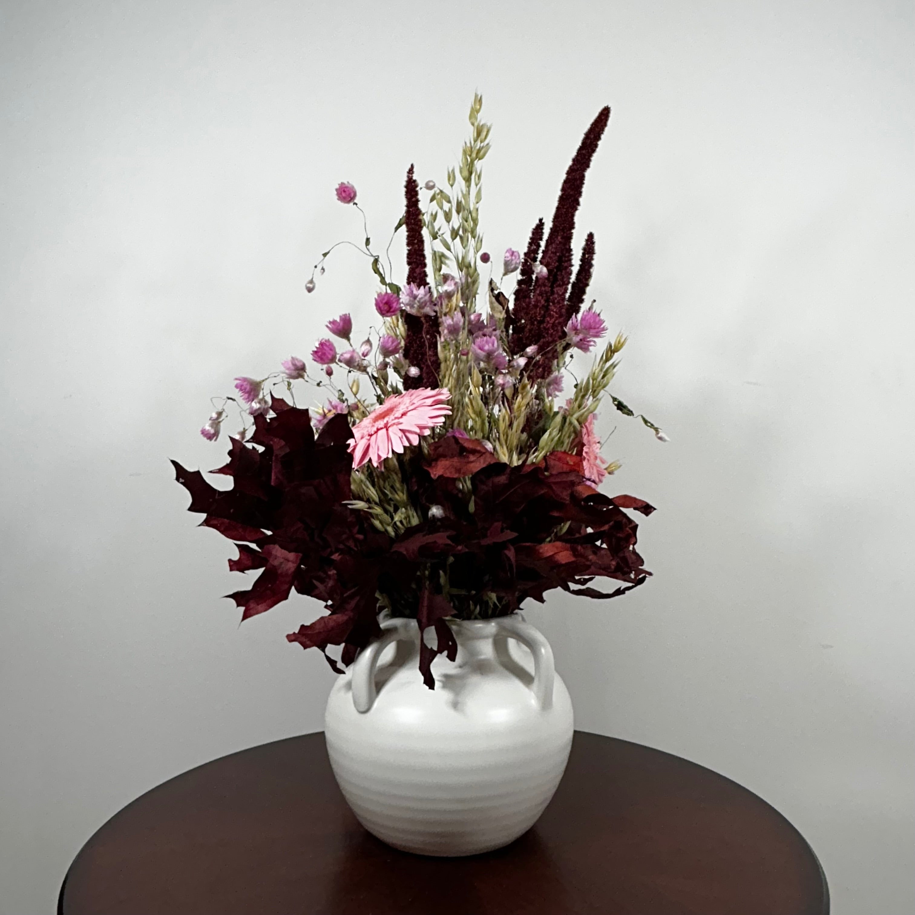 A dried flower arrangement in a white ceramic vase with two handles, set on a dark round table against a plain light gray wall.