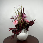 A dried flower arrangement in a white ribbed ceramic vase, displayed on a dark brown round table against a plain light gray wall.