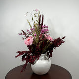 A dried flower arrangement in a white ribbed ceramic vase, displayed on a dark brown round table against a plain light gray wall.
