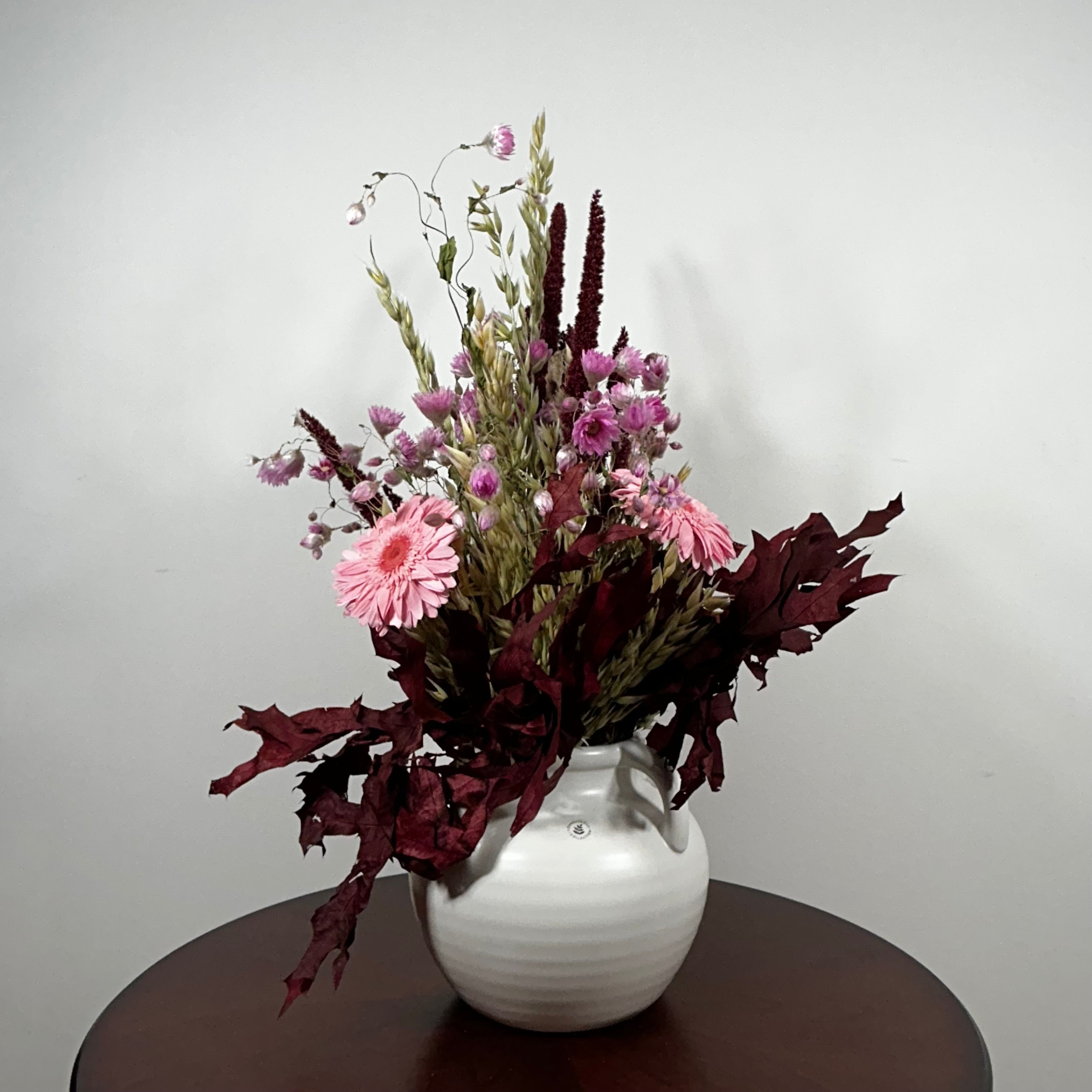 A dried flower arrangement in a white ribbed ceramic vase, displayed on a dark brown round table against a plain light gray wall.