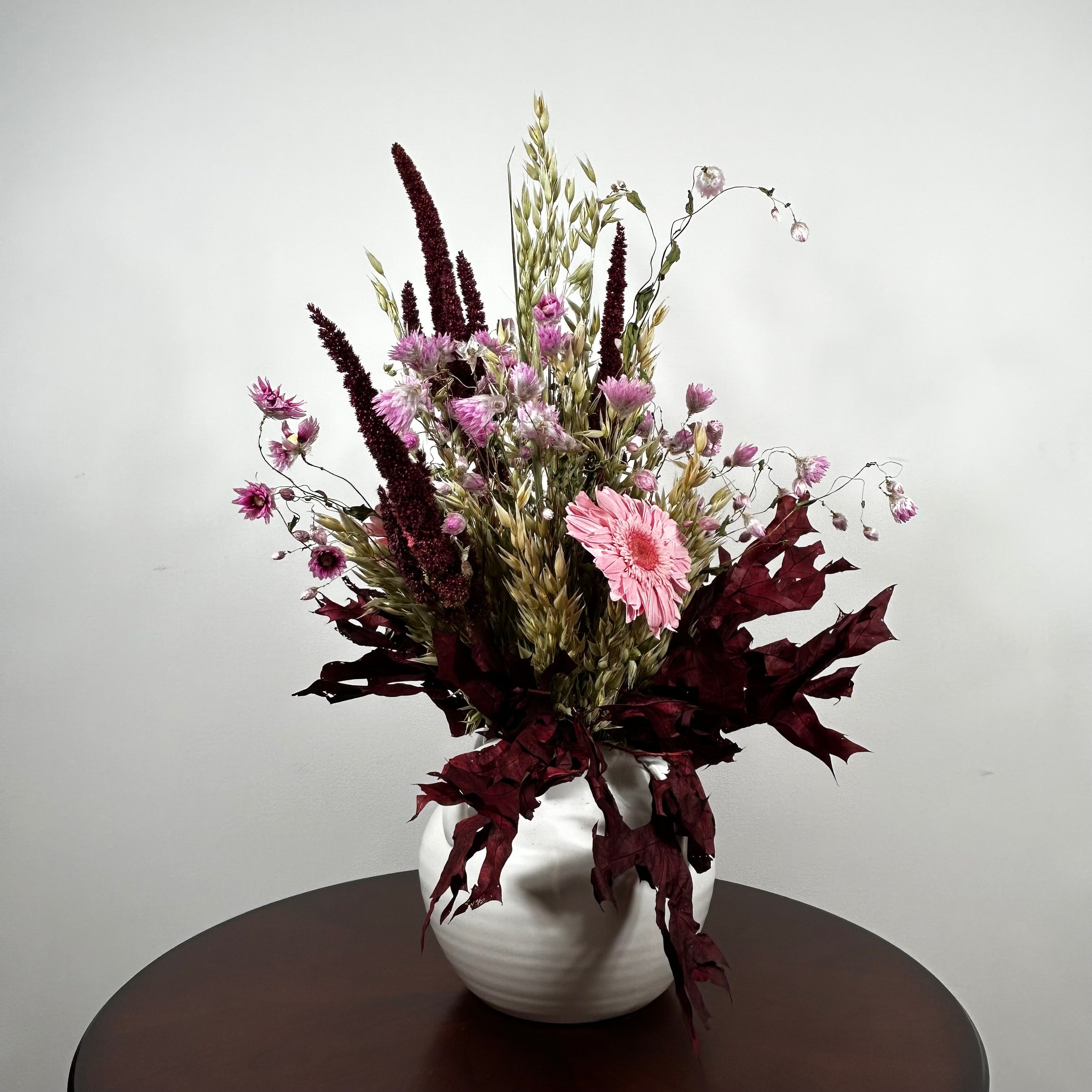 A dried floral arrangement in a white ceramic vase, featuring pink flowers, dark burgundy foliage, and tan grasses, displayed on a dark round table against a light gray wall.