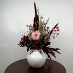 A dried floral arrangement in a white ceramic vase with two side handles, placed on a dark round table against a light gray wall. The arrangement features a light pink gerbera daisy, small pink flowers, burgundy and dark red foliage, and tan grasses.