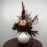 A dried floral arrangement in a white ceramic vase with two side handles, placed on a dark round table against a light gray wall. The arrangement features a light pink gerbera daisy, small pink flowers, burgundy and dark red foliage, and tan grasses.