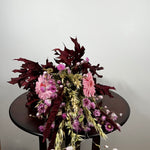 A dried floral arrangement featuring pink gerbera daisies, burgundy maple leaves, and pink amaryllis mixed with dried wheat stalks, displayed on a round dark wooden table against a plain light gray wall.