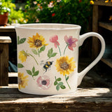 White mug with floral and bee design on a wooden surface with garden background