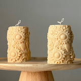 Two intricately designed candles embossed with sunflowers on a wooden stand with a grey background