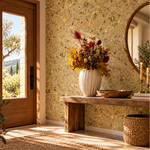 A styled interior entryway with a rustic wooden bench, a white ribbed ceramic vase filled with autumn flowers, a round mirror, and a woven basket. The wall features Alpine Meadow wallpaper in warm beige, yellow, and brown tones, and a wooden door with a glass window stands to the left. 3