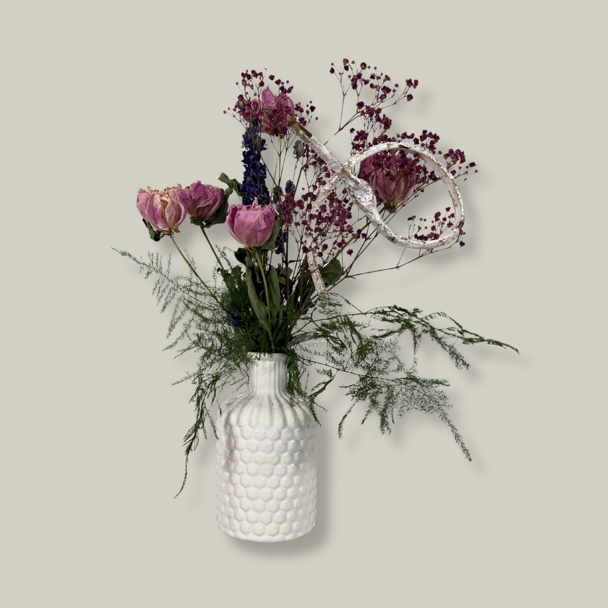 A modern white ceramic vase holds a dried flower arrangement with a delicate base of preserved asparagus ferns, from which soft plumes of gypsophila in a vibrant magenta, purple Larkspur, and soft pink peonies emerge. A final touch of playful charm is added by a single, beautifully sculpted white washed curly banana stick