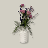 A modern white ceramic vase holds a dried flower arrangement with a delicate base of preserved asparagus ferns, from which soft plumes of gypsophila in a vibrant magenta, purple Larkspur, and soft pink peonies emerge. A final touch of playful charm is added by a single, beautifully sculpted white washed curly banana stick