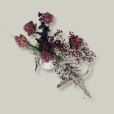 Top view of an arrangement with a delicate base of preserved asparagus ferns, from which soft plumes of gypsophila in a vibrant magenta, purple Larkspur, and soft pink peonies emerge. A final touch of playful charm is added by a single, beautifully sculpted white washed curly banana stick.