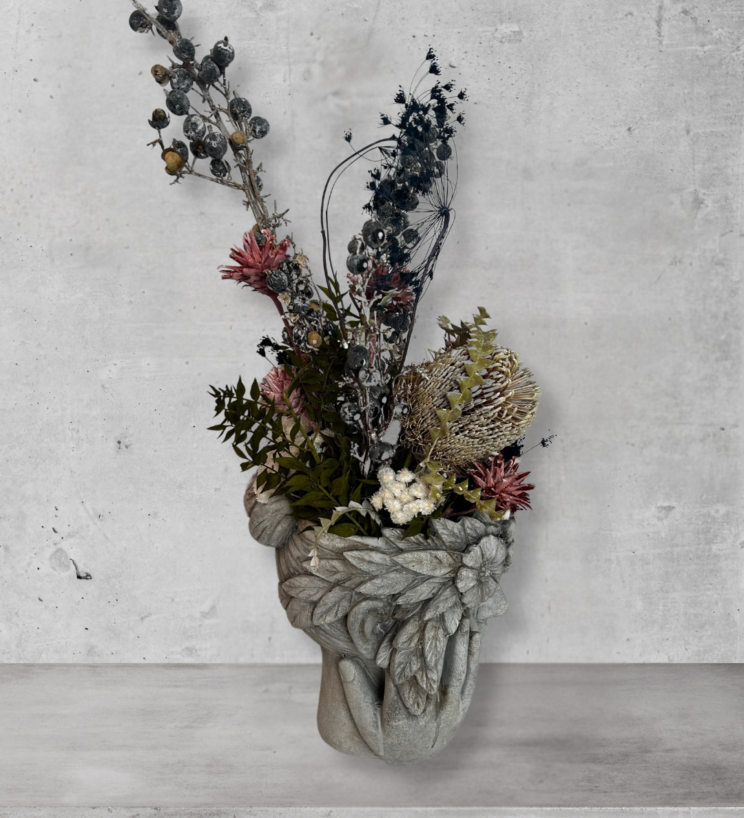 Side view of a decorative wall vase shaped like a face with hands on cheeks with dried flowers against a concrete wall.