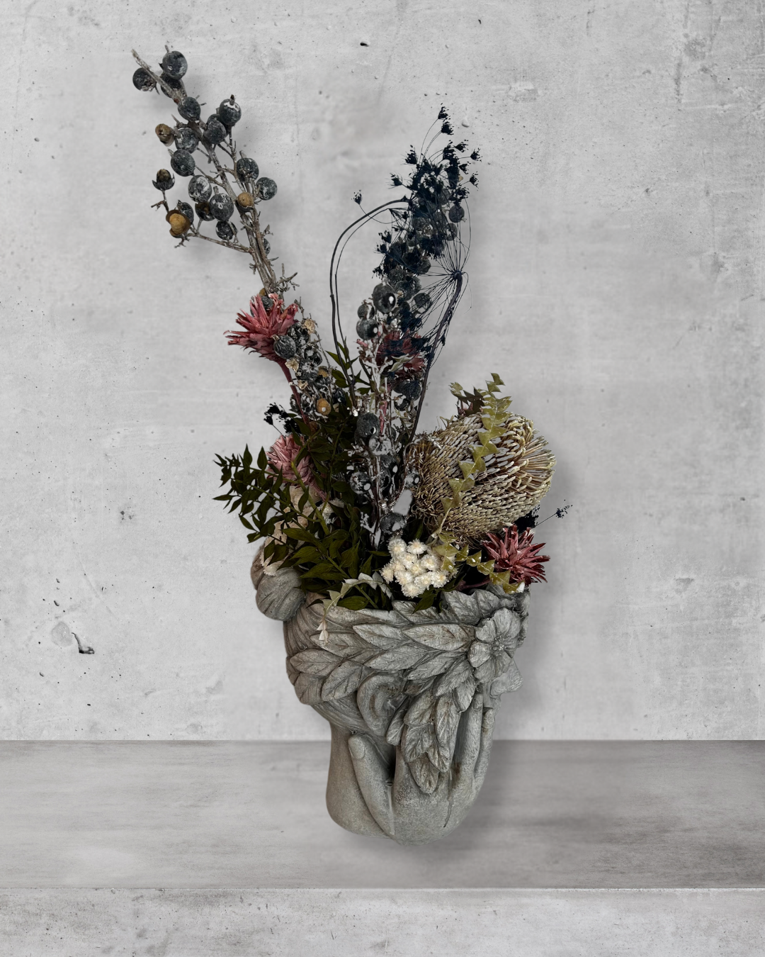 Side view of a decorative wall vase shaped like a face with hands on cheeks with dried flowers against a concrete wall.