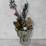 Side view of a decorative wall vase shaped like a face with hands on cheeks with dried flowers against a concrete wall.
