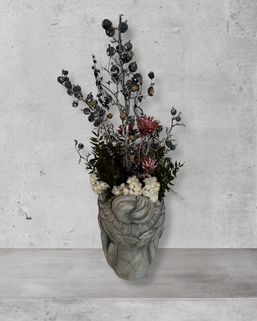 Back view of a decorative wall vase shaped like a face with hands on cheeks with dried flowers against a concrete wal