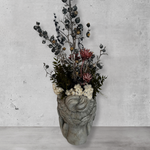 Back view of a decorative wall vase shaped like a face with hands on cheeks with dried flowers against a concrete wal