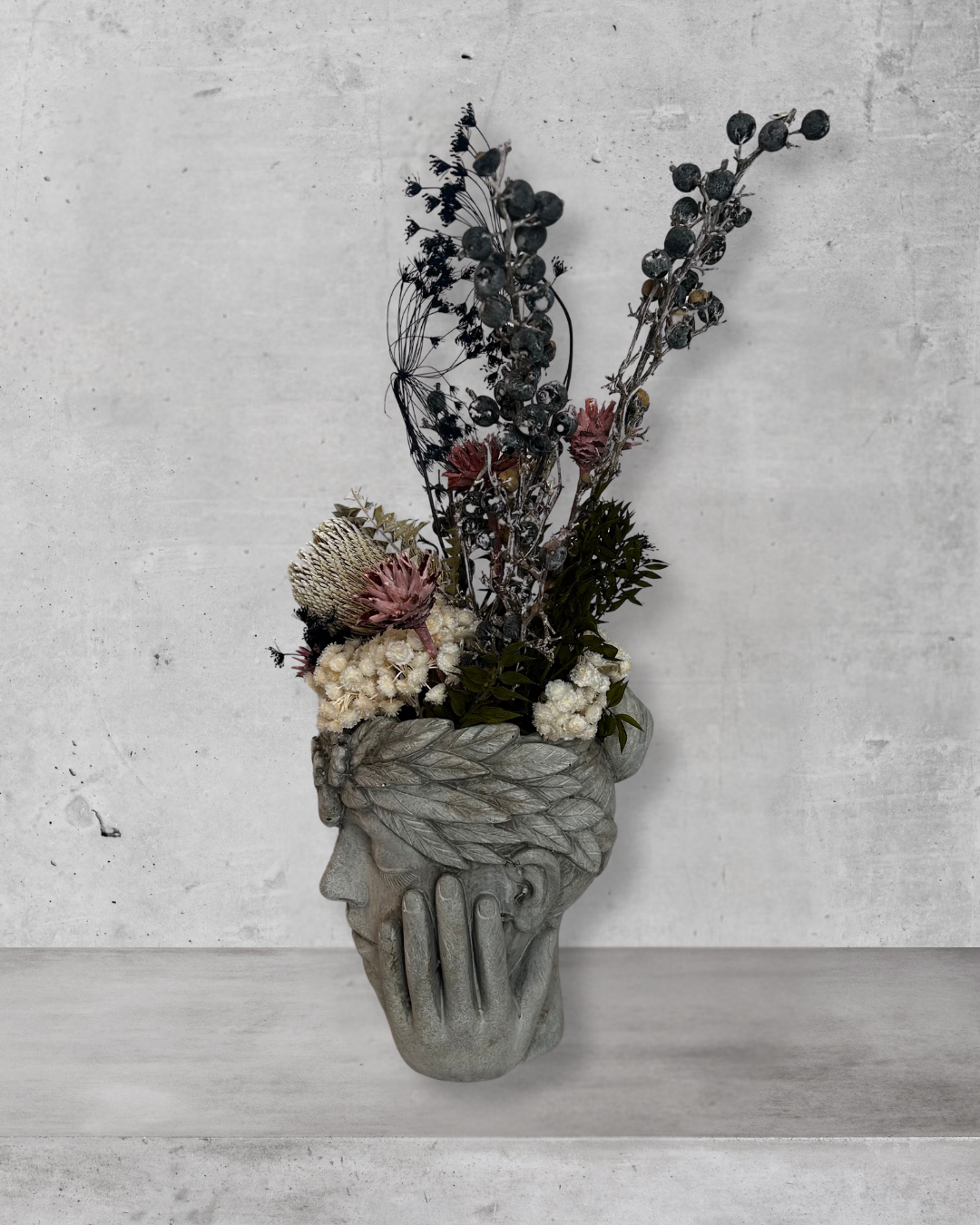 Side view of a decorative wall vase shaped like a face with hands on cheeks with dried flowers against a concrete wal