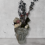 Side view of a decorative wall vase shaped like a face with hands on cheeks with dried flowers against a concrete wal
