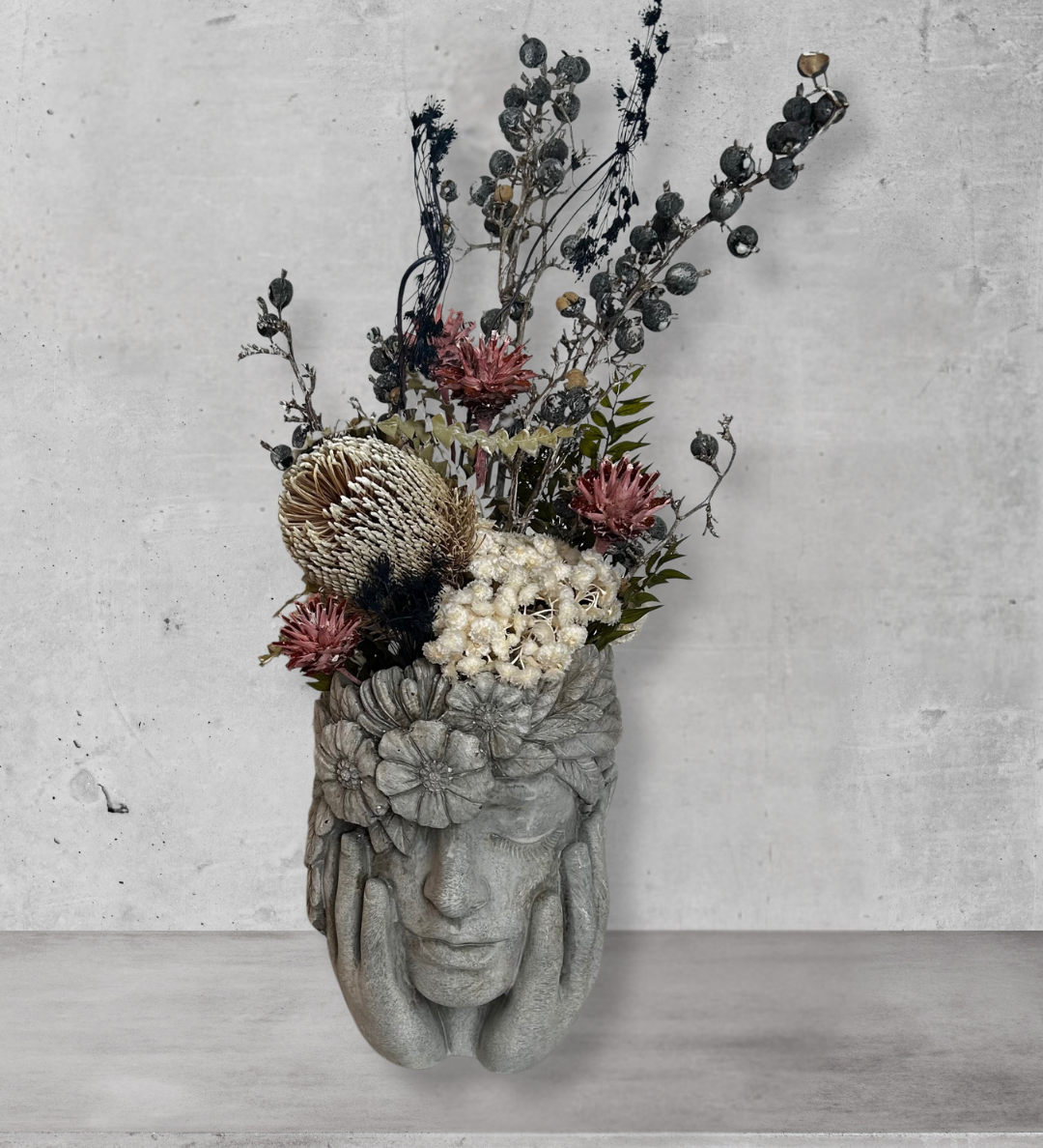 Decorative wall vase shaped like a face with hands on cheeks with dried flowers against a concrete wall.