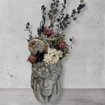 Decorative wall vase shaped like a face with hands on cheeks with dried flowers against a concrete wall.
