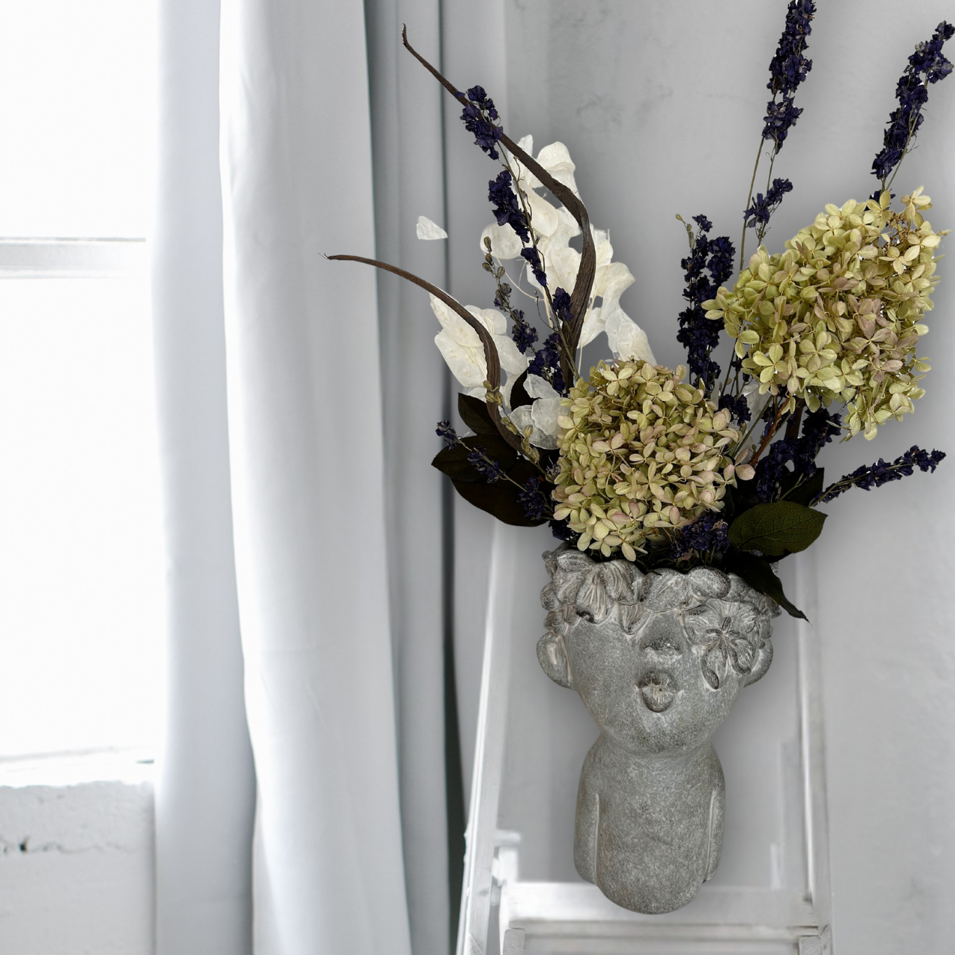 Decorative everlasting flower arrangement in a concrete vase sculpted as a woman's serene face with her lips charmingly puckered, on a white chair against a white curtain background