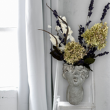 Decorative everlasting flower arrangement in a concrete vase sculpted as a woman's serene face with her lips charmingly puckered, on a white chair against a white curtain background