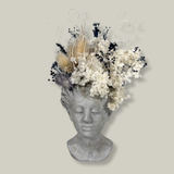 This concrete vessel, beautifully sculpted as a woman's serene face, her hair appears to be caught in a soft breeze, with a dried floral arrangement made up navy bupleurum , white curly ting ting, delicate light blue cape flowers, a cluster of three bleached teasels adorns the front left side of her face, and a pure white hydrangea on the right. 