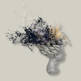 Side view of a concrete vessel, beautifully sculpted as a woman's serene face, her hair appears to be caught in a soft breeze, with a dried floral arrangement made up navy bupleurum , white curly ting ting, delicate light blue cape flowers, and a cluster of three bleached teasels. 