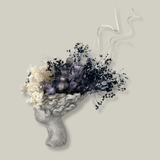 Side view of a concrete vessel, beautifully sculpted as a woman's serene face, her hair appears to be caught in a soft breeze, with a dried floral arrangement made up navy bupleurum , white curly ting ting, delicate light blue cape flowers, and a pure white hydrangea. 