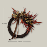 Decorative asymmetrical wreath that blends rustic textures with rich autumnal colours, accented with a rich brown velvet ribbon hung on a wall, showing measurements of 16 inches wide by 22 inches tall.