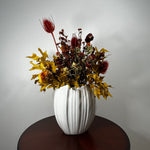 A white ribbed ceramic vase filled with an autumnal dried floral arrangement featuring yellow maple leaves, red seed pods, and dark burgundy botanicals, displayed on a dark wooden table against a light gray wall.