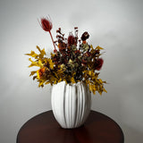 A white ribbed ceramic vase filled with an autumnal dried floral arrangement featuring yellow maple leaves, red seed pods, and dark burgundy botanicals, displayed on a dark wooden table against a light gray wall.