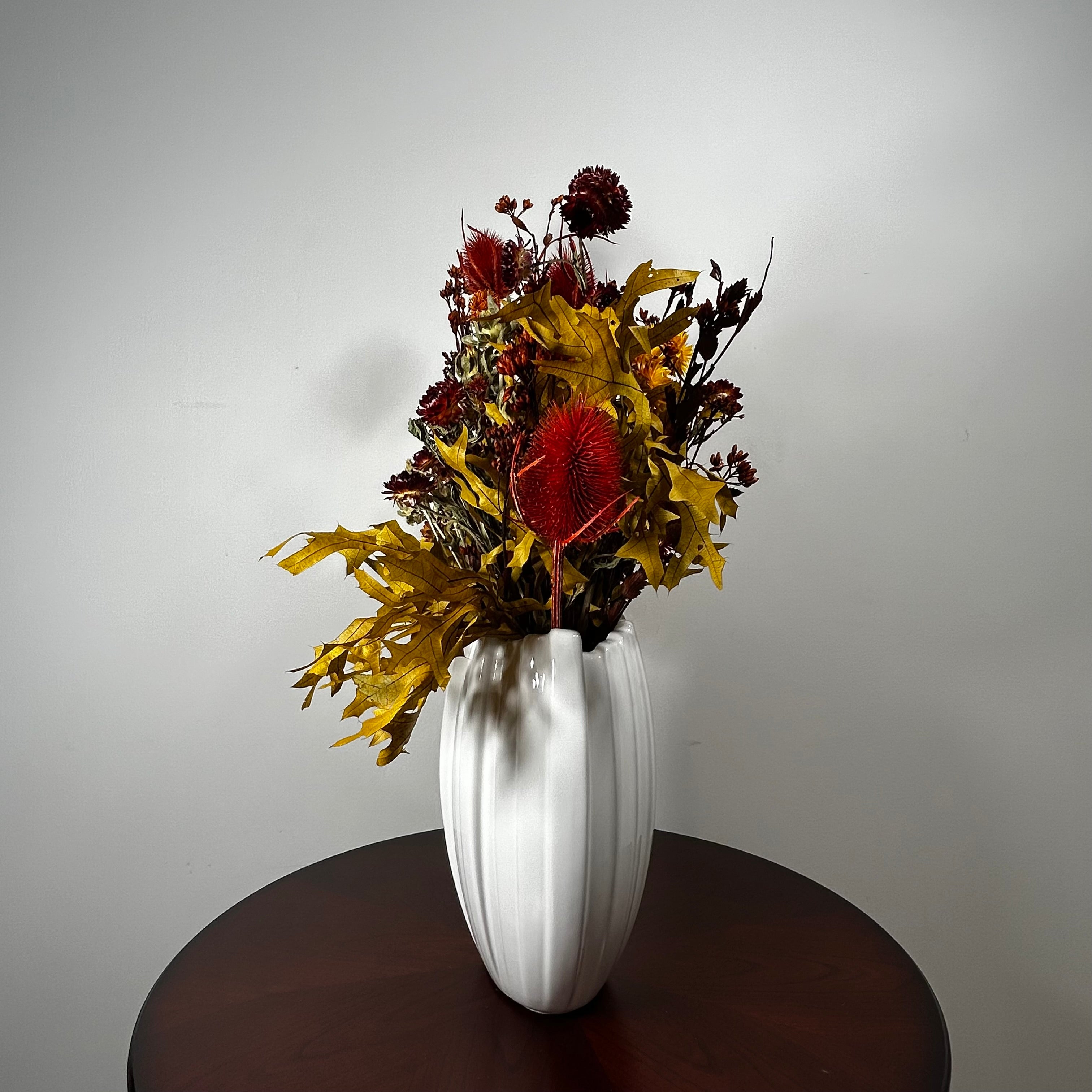 A white ribbed ceramic vase filled with a dried floral arrangement featuring red and yellow botanicals, displayed on a dark wooden table against a plain light gray wall.