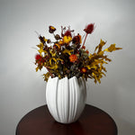 A white ribbed ceramic vase filled with an autumnal dried flower arrangement, featuring golden maple leaves, red thistle-like blooms, and yellow-orange flowers, displayed on a dark round wooden table against a light gray wall.