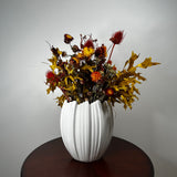 A white ribbed ceramic vase filled with an autumnal dried flower arrangement, featuring golden maple leaves, red thistle-like blooms, and yellow-orange flowers, displayed on a dark round wooden table against a light gray wall.
