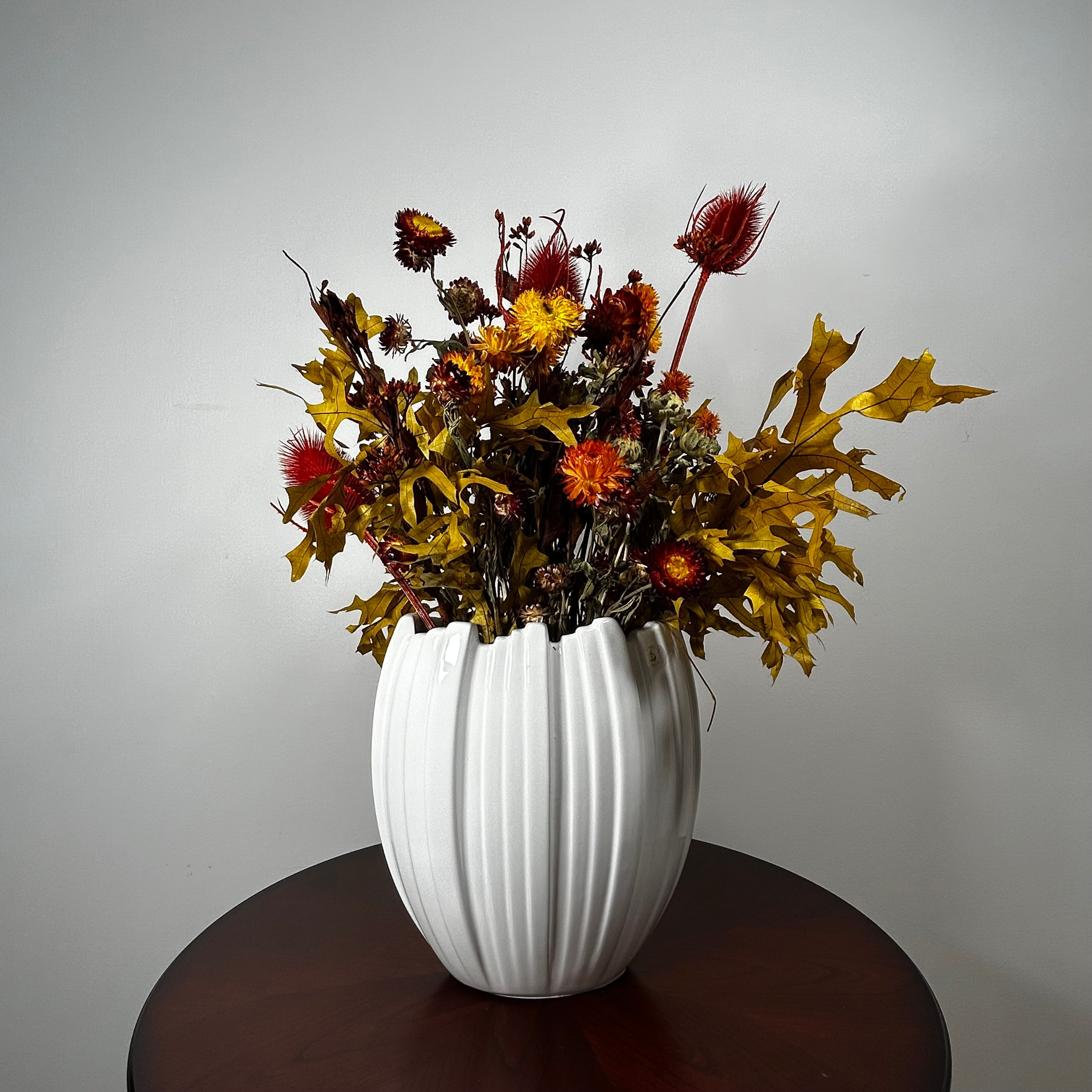 A white ribbed ceramic vase filled with an autumnal dried flower arrangement, featuring golden maple leaves, red thistle-like blooms, and yellow-orange flowers, displayed on a dark round wooden table against a light gray wall.
