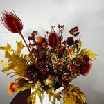 A dried floral arrangement in a white ceramic vase, featuring golden oak leaves, red feathery stems, and mixed autumn flowers against a plain light gray wall.